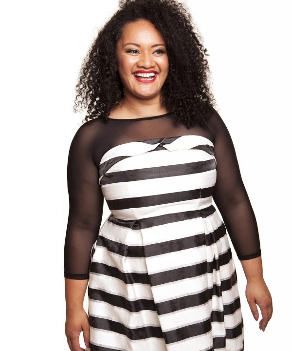 Vibrant woman smiling in black and white striped dress with sheer black sleeves, joyful expression, curly hair, modern fashion, bright studio portrait, confident and stylish.