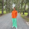 Vibrant young man standing on a gravel pathway surrounded by tall green trees, wearing a bright orange shirt and mint green pants, embodying cheerful and colorful fashion in a natural outdoor setting.