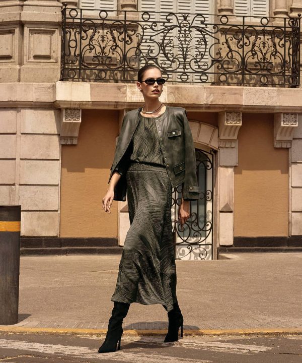 Stylish woman model wearing a chic outfit with a leather jacket, patterned dress, and high-heeled boots, standing on city street for fashion photography.
