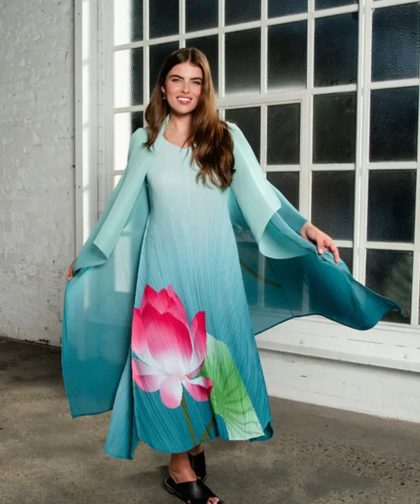 Elegant woman wearing a flowy, colorful dress with a large pink floral print, standing indoors near a large industrial-style window. Perfect for fashion, spring collection, or floral-inspired designs.