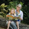 Reading book with grandmother and granddaughter outdoors in nature, promoting family bonding, literacy, and outdoor activities, sunny day, lush green background, children’s storybook, learning, nature exploration, bonding with family, outdoor reading, educational activities, storytime.