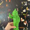 Colorful green resin sculpture of a quirky man with a raised middle finger, set against an abstract textured black background with splashes of red, yellow, and white paint.