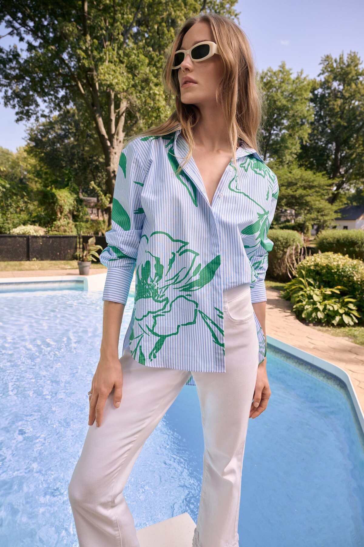 1. Stylish woman wearing a blue and green floral print shirt, white pants, and eco-friendly sunglasses by the poolside on a sunny day, showcasing fashion from katyamaker.